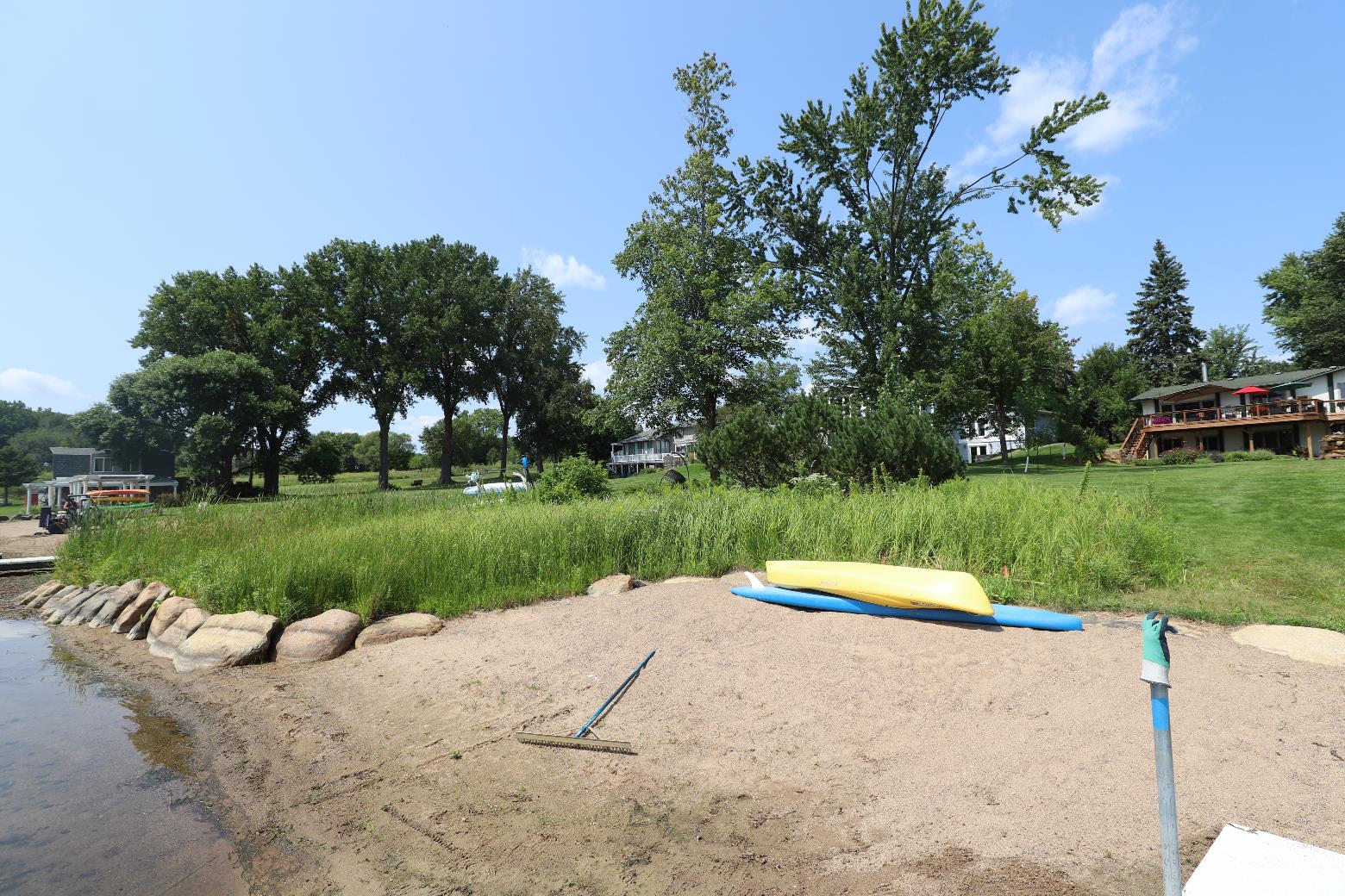 A sandy beach with a rake lies by a lake. Two kayaks, one yellow and one blue, rest on the shore. Trees and grass lead up to several houses in the background under a clear blue sky